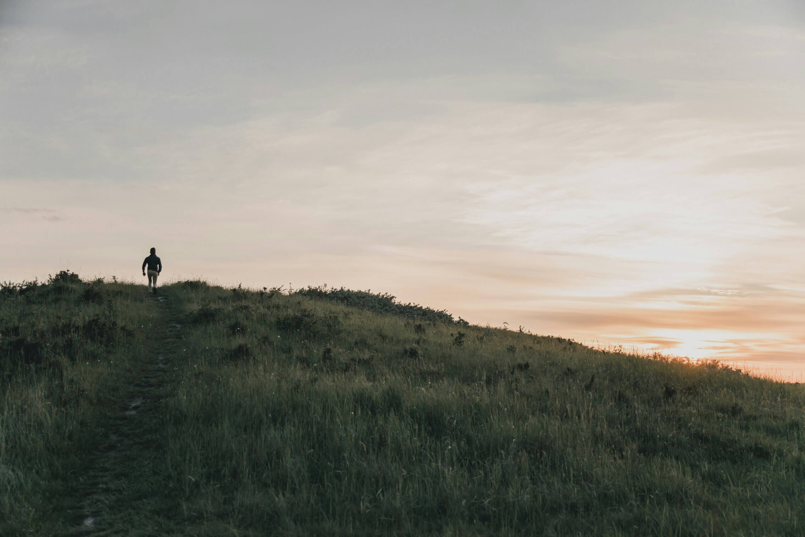 A person walks alone along a grassy hill at sunset, silhouetted against a soft sky with warm orange light near the horizon.